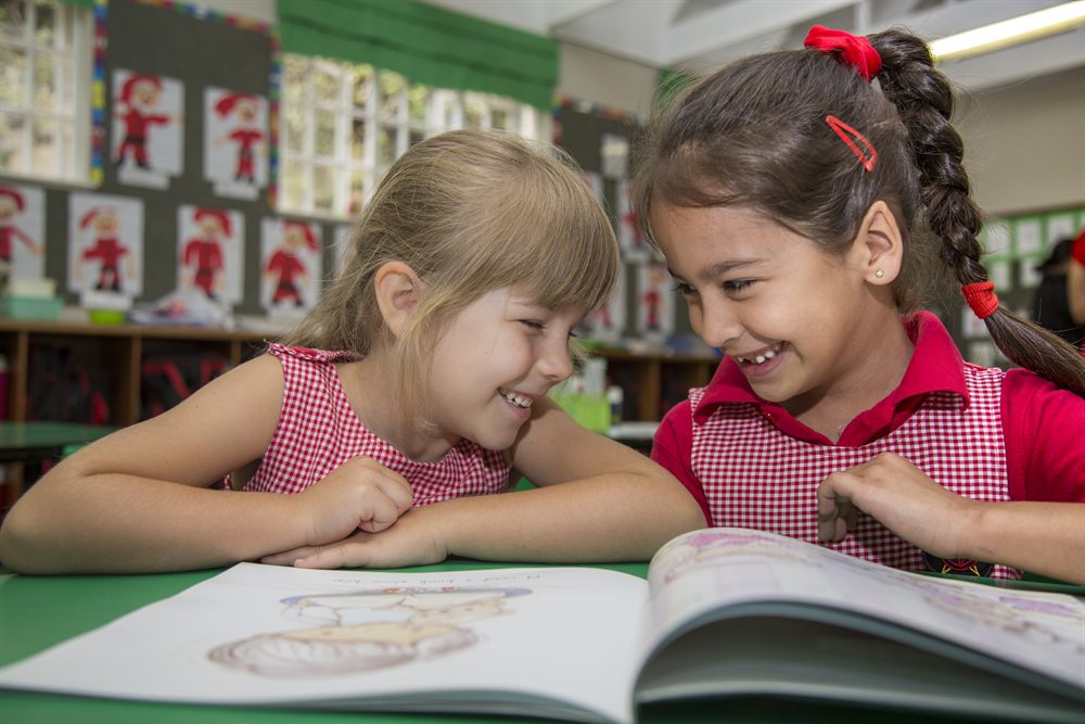 Two Little Anchors girls in pink gingham laughing over a picture book
